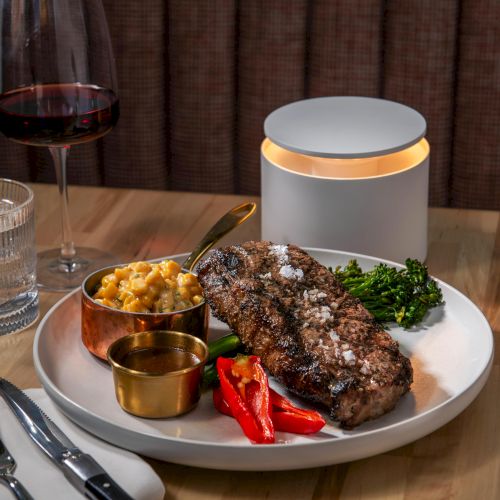 A steak dinner with vegetables, macaroni and cheese, a candle, and glasses of water and red wine on a wooden table.