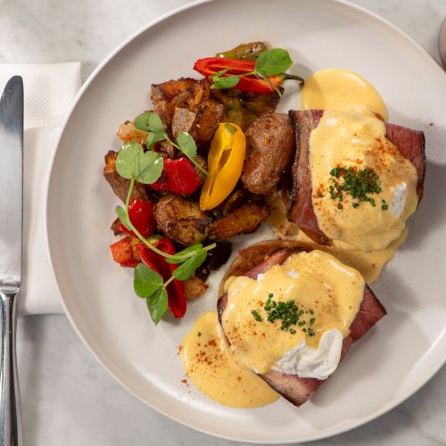 The image shows eggs Benedict with hollandaise sauce, roasted potatoes with peppers, a fork, knife, white napkin, and a drink nearby.