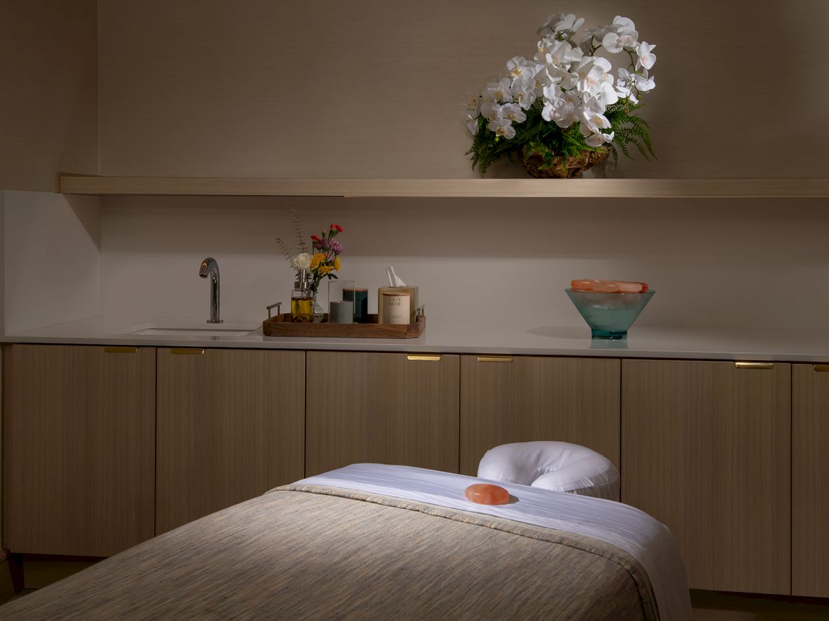A serene spa room with a massage table, orchids on a shelf, a water tap, and decorative items, creating a calming atmosphere.