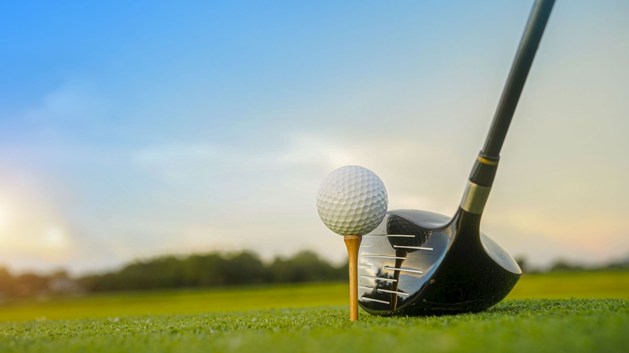 A golf ball on a wooden tee is positioned next to a driver club on a lush green course, against a clear sky.