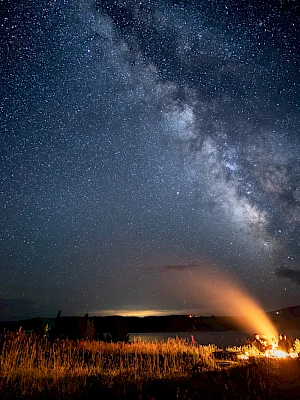 A campfire glows under a starry sky with the Milky Way visible, surrounded by an open field and distant horizon.