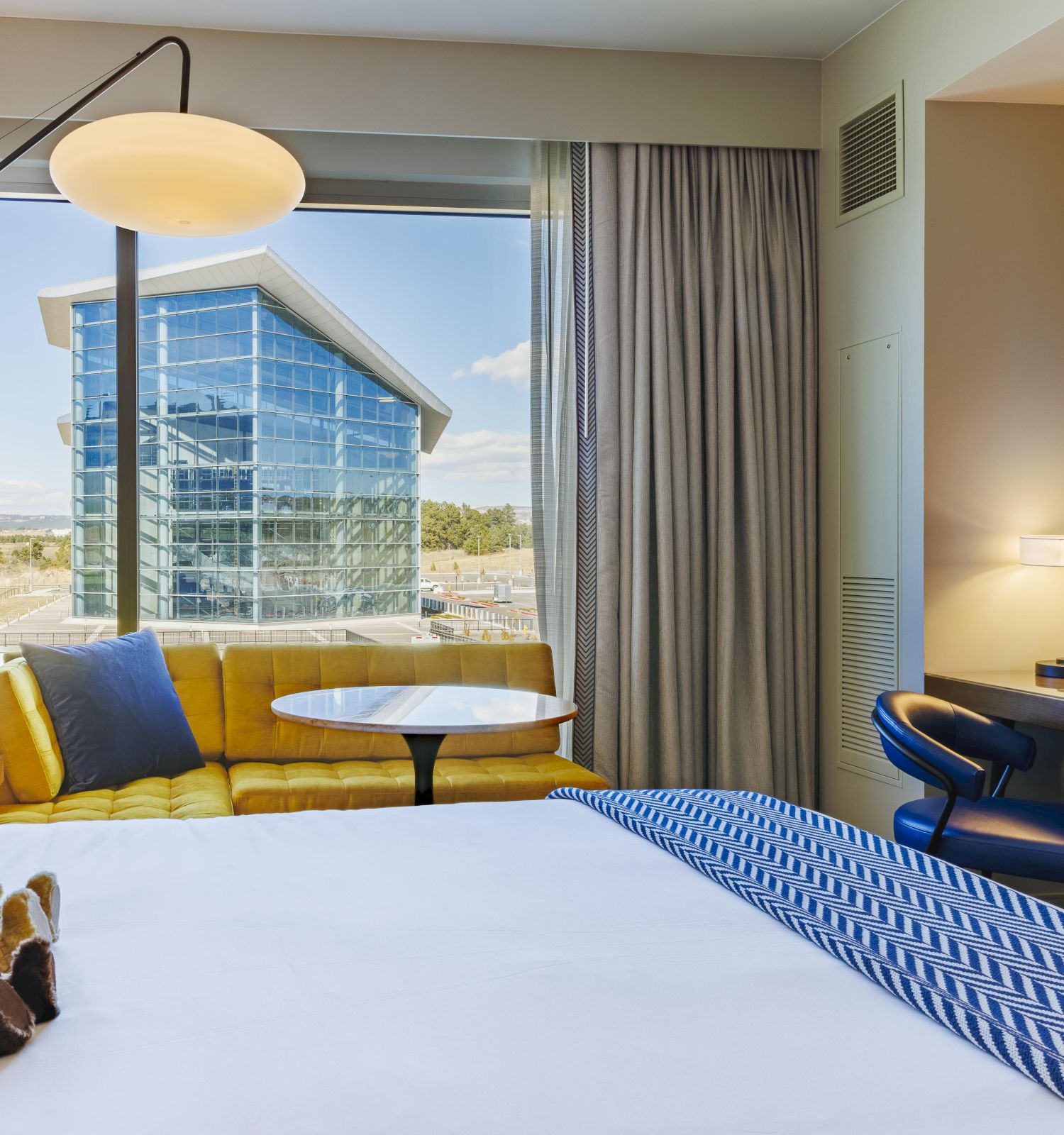 A modern hotel room features a bed with a teddy bear, a yellow couch, a desk, and a window view of a contemporary building.