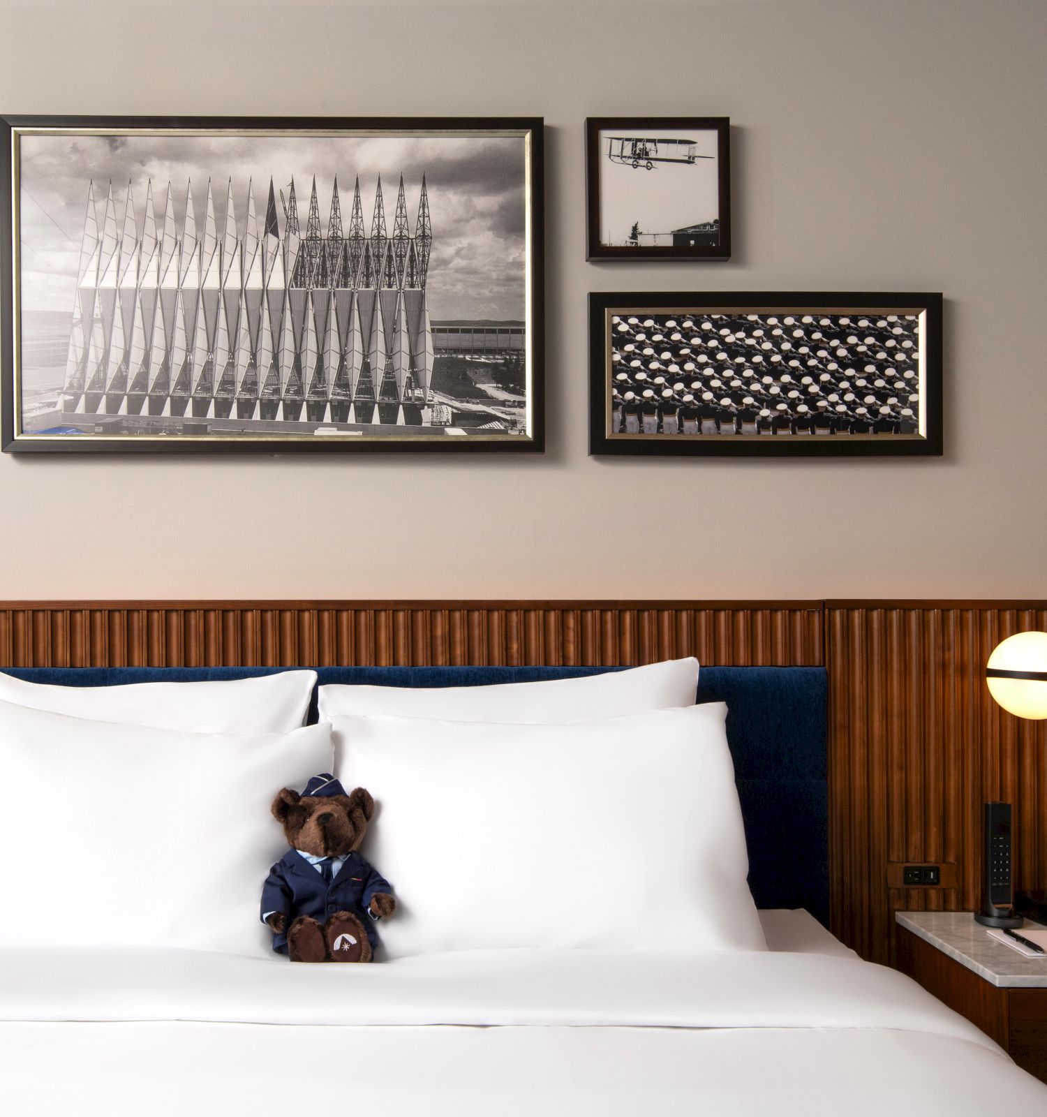A neatly made bed with a teddy bear, modern lamps, and framed photos on the wall above.