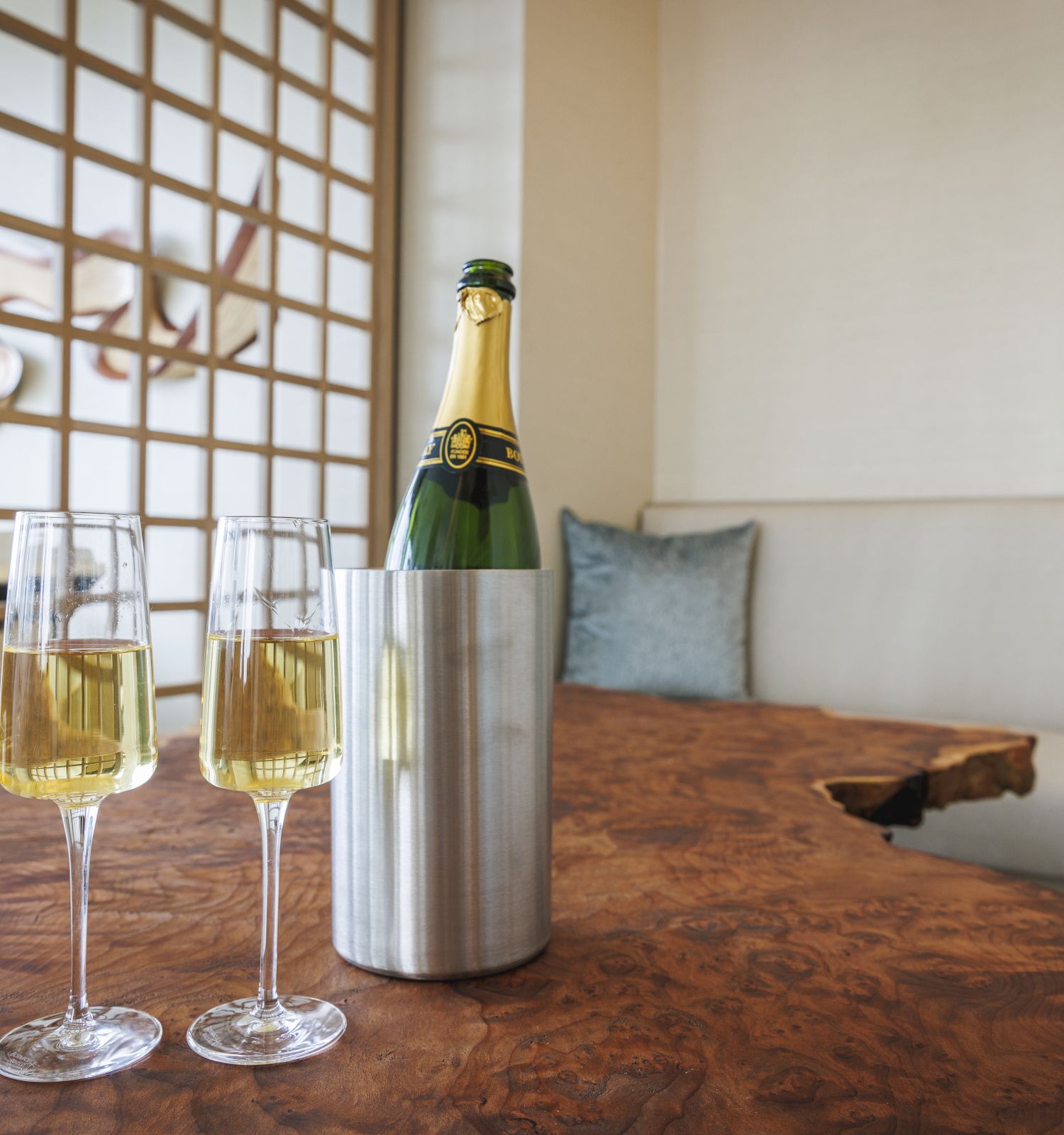 A bottle of champagne in a cooler with two filled glasses on a wooden table in a cozy room setting.