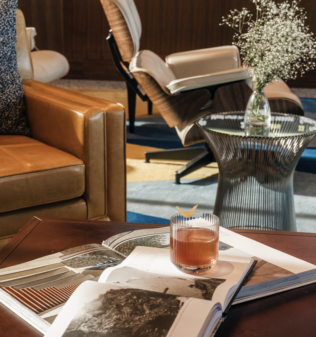 Cozy lounge with a leather chair, open book, a glass of drink, and a modern side table; plant bouquet adds calm ambiance.