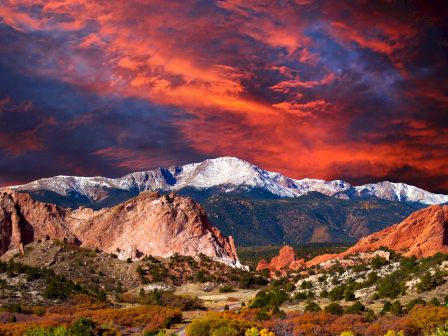 A stunning landscape with red rock formations, a snow-capped mountain, and a dramatic, colorful sky at sunset.