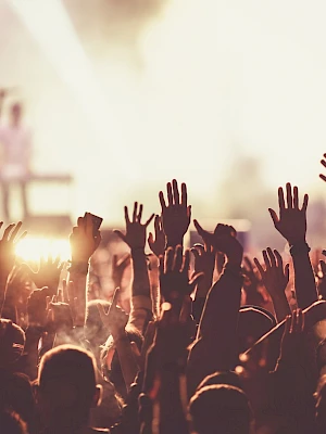 A crowd with raised hands at a concert, with bright lights and performers blurred in the background, creates a lively atmosphere.