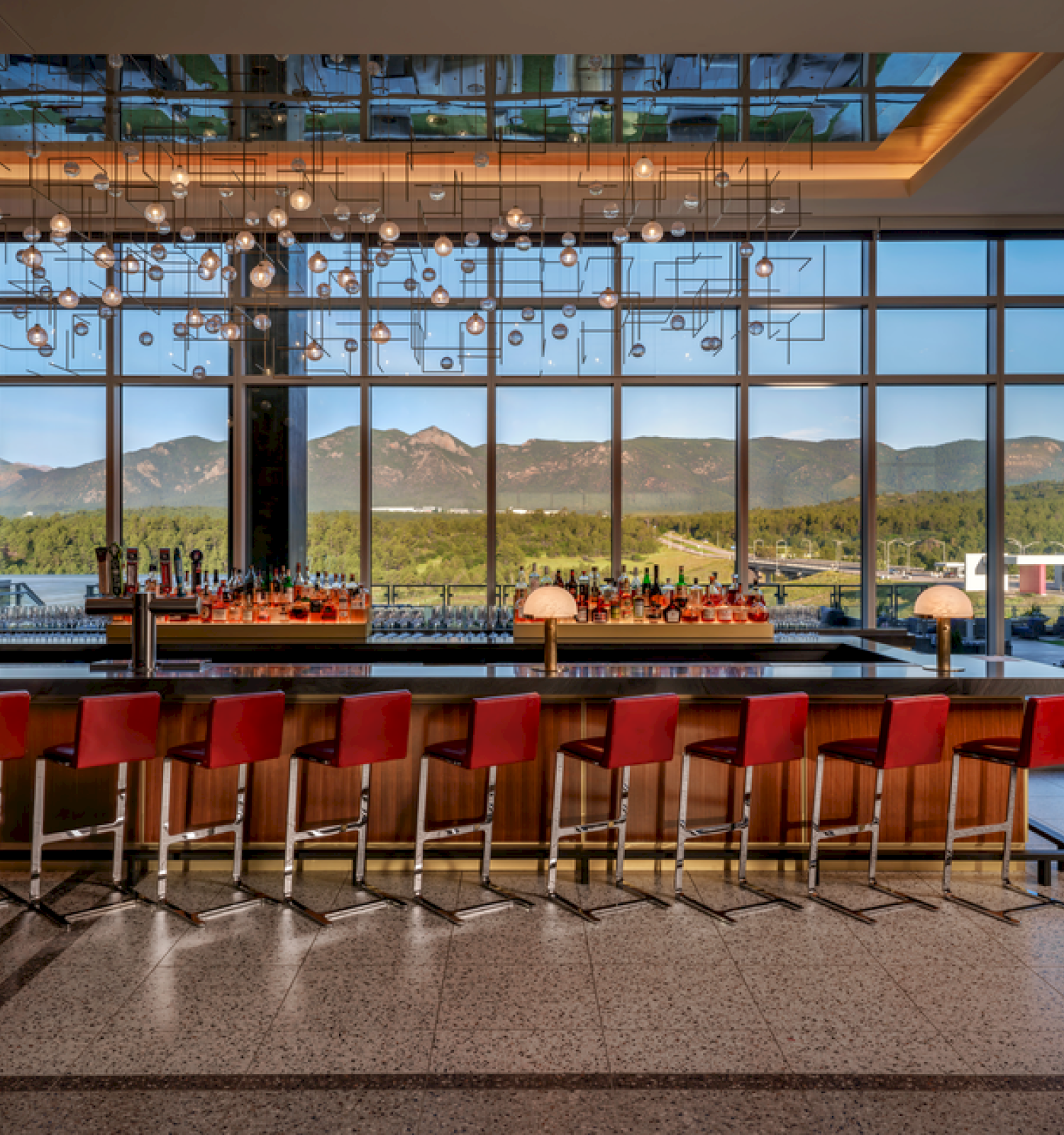 A modern bar with red stools and a chandelier, offering a scenic view of mountains through large windows.