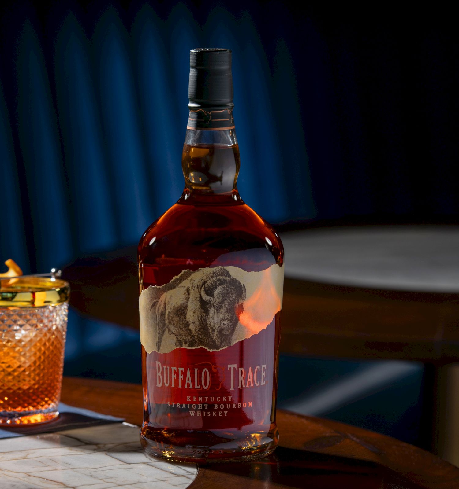 A bottle of Buffalo Trace whiskey and a cocktail glass with an orange garnish are on a table.