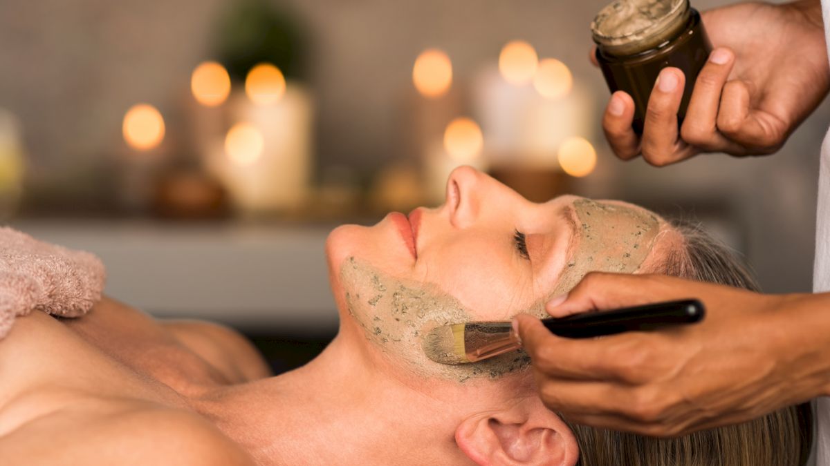 A person receiving a facial treatment, with a brush applying a mask, candles blurred in the background creating a relaxing ambiance.