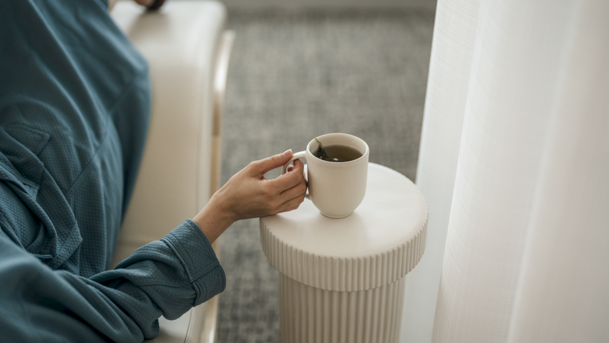 A person in a blue robe lounges on a sofa, holding a mug on a small table by a window with sheer curtains.