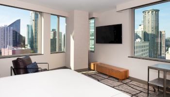 The image shows a modern hotel room with a large bed, a chair, a wall-mounted TV, and large windows offering a city skyline view.