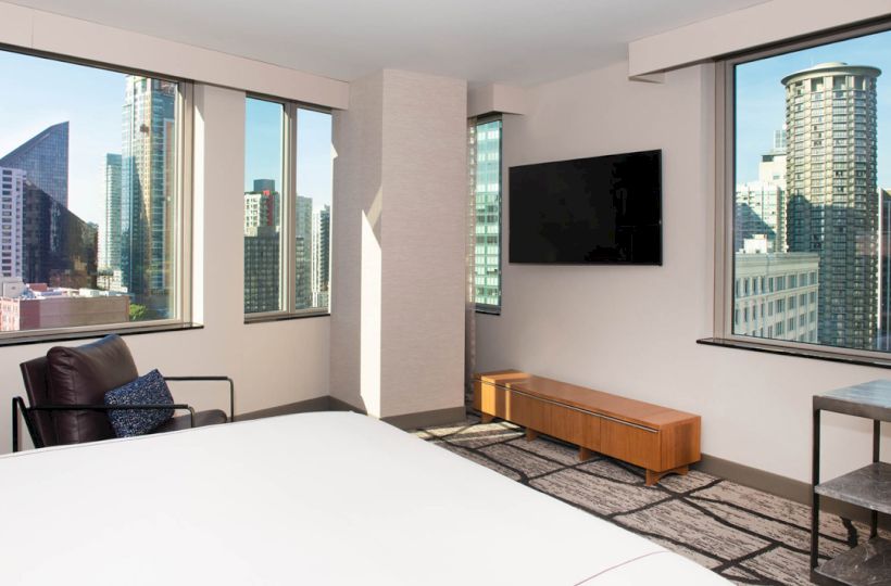 The image shows a modern hotel room with a large bed, a chair, a wall-mounted TV, and large windows offering a city skyline view.