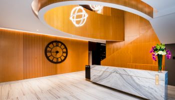 The image shows a modern lobby with a marble reception desk, a large clock on the wall, contemporary lighting fixtures, and a bouquet of flowers.