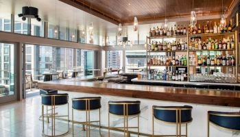 A modern, stylish bar with a well-stocked liquor shelf, blue and gold bar stools, and large windows providing a city view in the background.