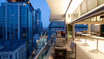 A modern balcony with tables and chairs overlooks a street lined with tall buildings during dusk, featuring a cityscape with lights on.