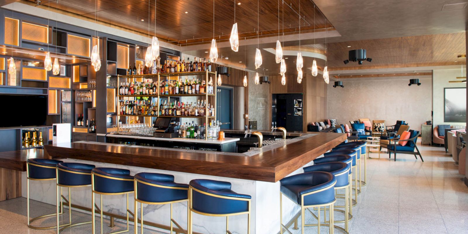 This image shows a modern bar with a large countertop, several bar stools, hanging lights, and a well-stocked selection of bottles in the background.