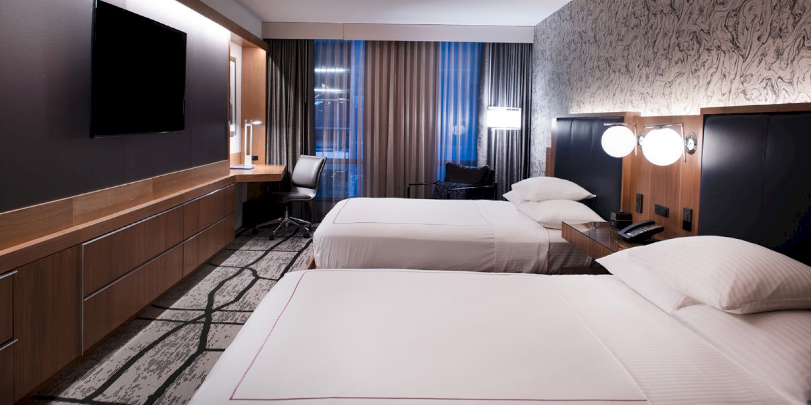 The image shows a modern hotel room with two neatly made beds, wall-mounted TV, desk, chair, nightstands with lamps, and patterned wallpaper.
