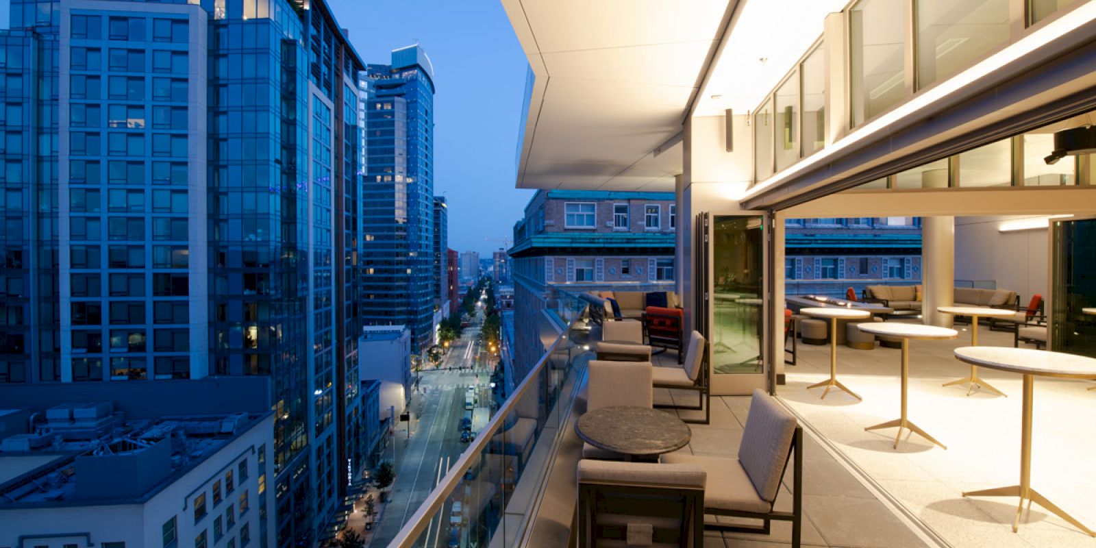 The image shows a modern cityscape viewed from a stylish, open balcony at dusk, featuring outdoor seating and tables.