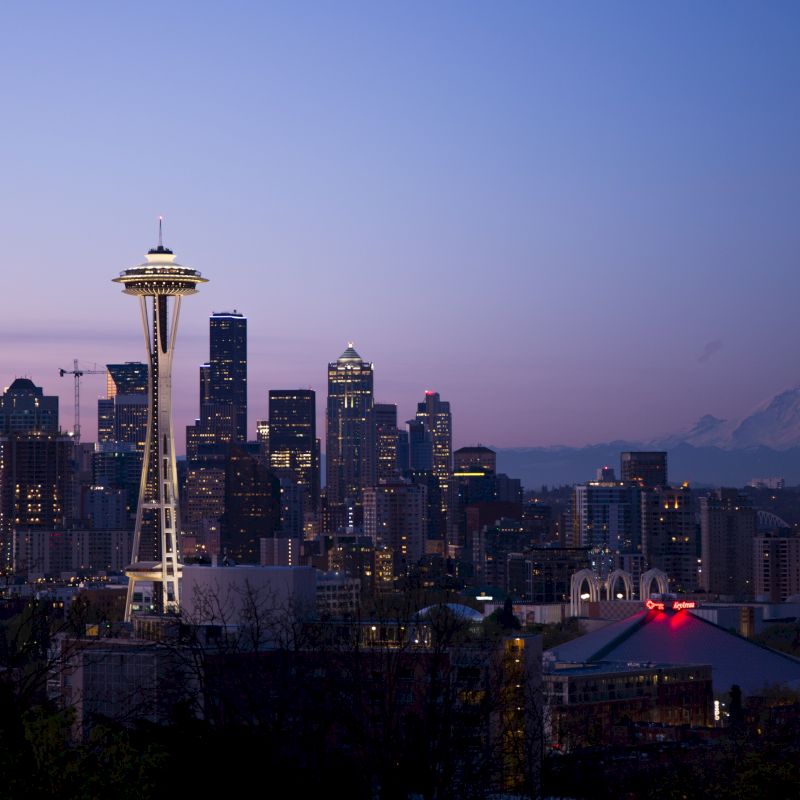 The image shows Seattle's skyline at dusk, featuring the Space Needle and Mount Rainier in the background, with city lights starting to illuminate.