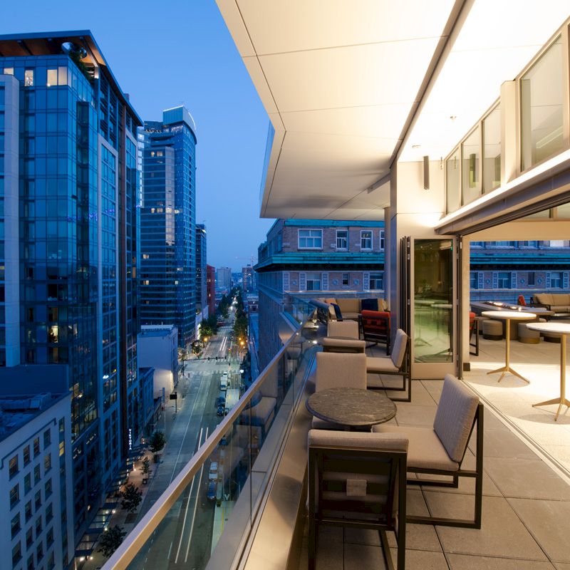 An outdoor balcony with seating is overlooking a cityscape at twilight, with modern buildings and illuminated streets below.