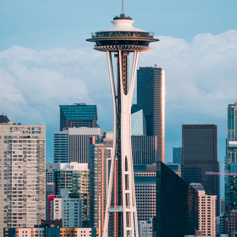 The image shows the Space Needle with a backdrop of skyscrapers and various buildings, set against a partially cloudy sky.