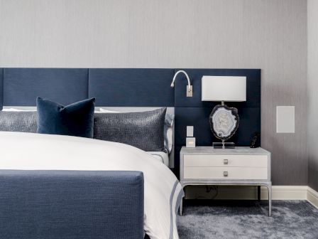 A modern bedroom with a navy blue headboard, white bedspread, and navy pillow. A nightstand holds a unique lamp and books beneath it, on a grey carpet.