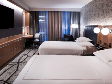 The image shows a modern hotel room with two neatly made beds, a flat-screen TV, a work desk, and large windows with curtains, ending the sentence.