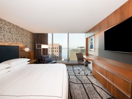 This image shows a modern hotel room with a king-sized bed, large TV, desk with chair, armchair, floor-to-ceiling windows, and city view outside.