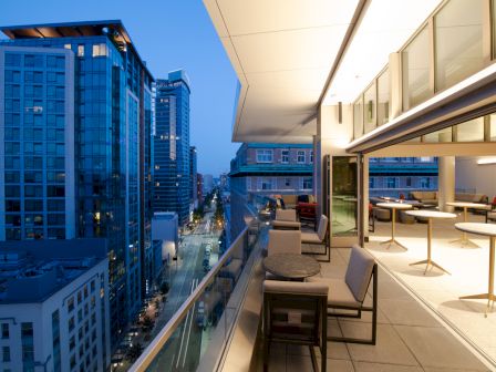 A modern cityscape at dusk with tall buildings alongside a furnished balcony, featuring tables and chairs. The balcony overlooks the street below.