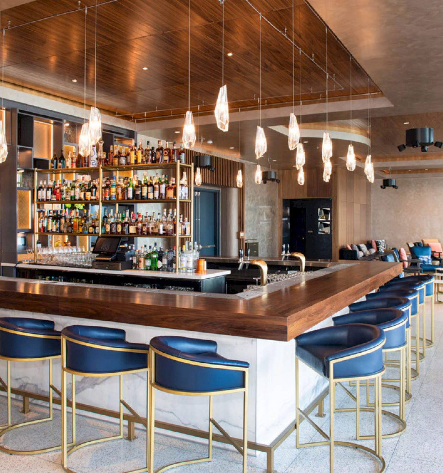 A modern bar with high stools, hanging lights, and a well-stocked liquor shelf. The seating area features sofas and tables in the background.