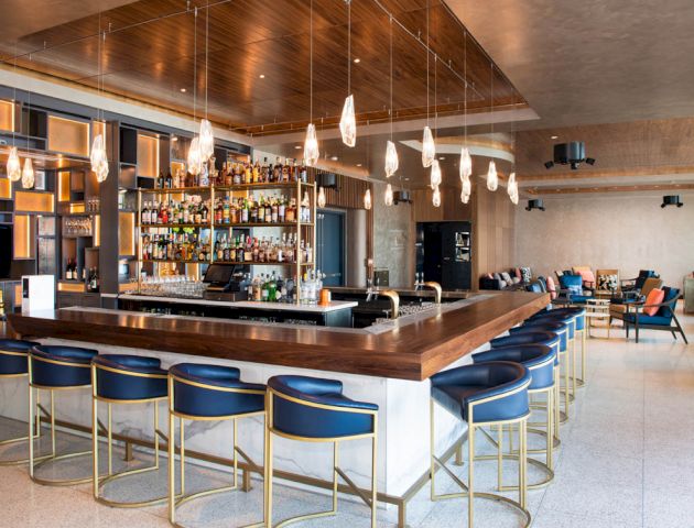 The image features a modern bar with blue barstools, hanging lights, and a stocked bar. There is seating and a lounge area in the background.
