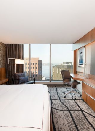 A modern hotel room with a large bed, desk, chair, flat-screen TV, and a window with a view of buildings and water outside.