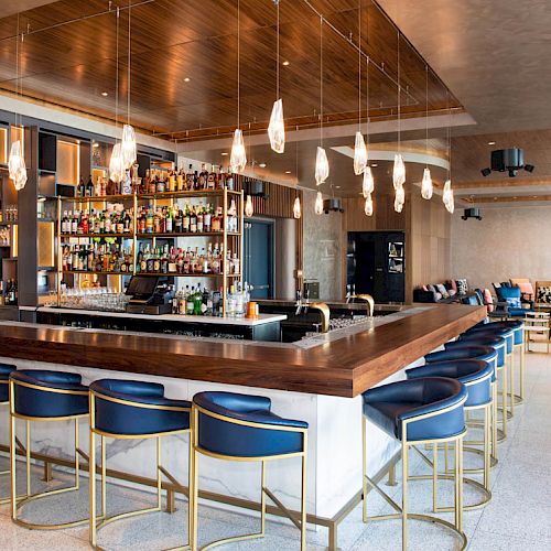 A modern bar with blue stools, wooden counters, a well-stocked liquor shelf, and elegant lighting hanging from the ceiling.