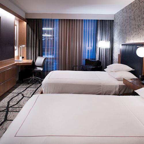 The image shows a modern hotel room with two neatly made beds, a wall-mounted TV, a work desk with a chair, and ambient lighting.