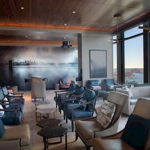 A modern lounge with ample seating, large windows offering a water view, and contemporary wall art of a city's skyline.