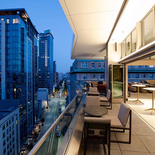 The image shows a modern urban balcony at dusk, overlooking a city street, with seating and tables. Buildings and streetlights are visible.
