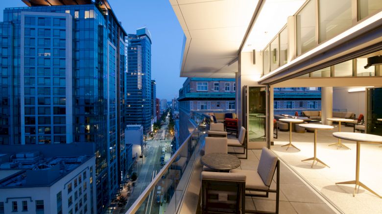 The image shows a modern cityscape at dusk with high-rise buildings and a well-lit balcony featuring tables and chairs on the right side.
