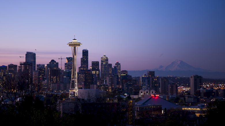 A city skyline at dusk with the iconic Space Needle prominently lit, surrounded by high-rise buildings, with mountains visible in the background.