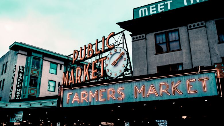 The image shows the iconic neon signs of a Public Market and Farmers Market, likely a famous marketplace, with surrounding buildings in the background.