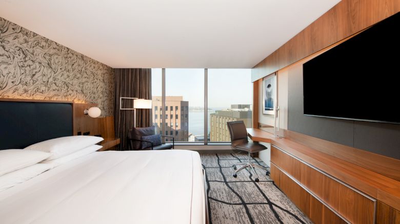 The image depicts a modern hotel room with a large bed, TV, desk, chair, and a window with a city view.