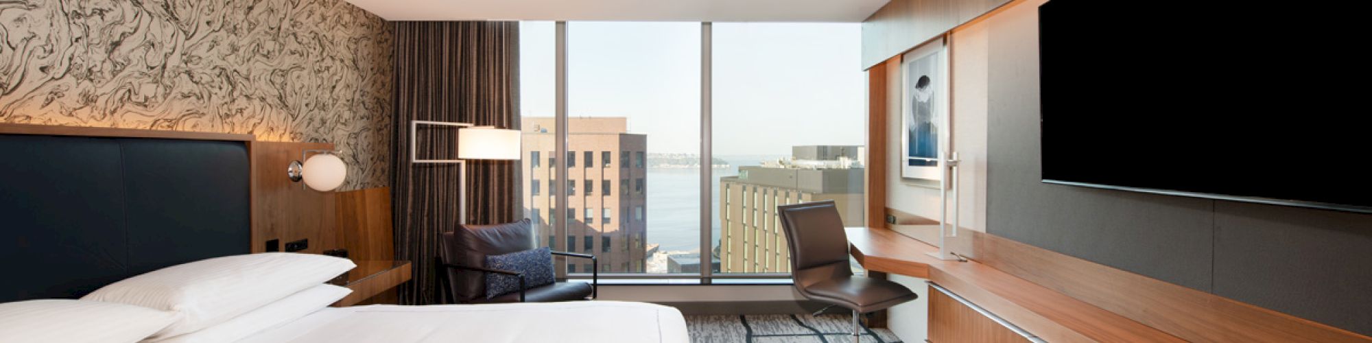 A modern hotel room with a large bed, desk, chair, wall-mounted TV, and a window with a city view. The decor is minimalistic and sleek.