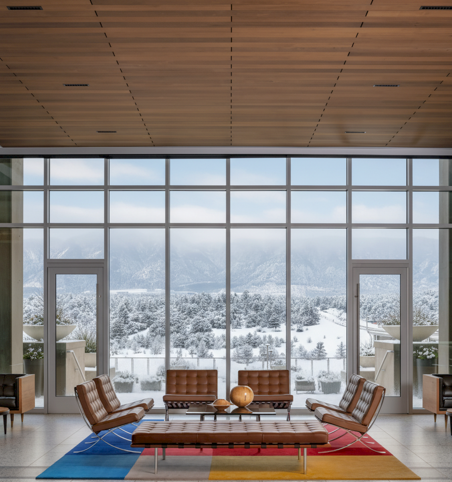 A modern lounge with chairs and a table on a colorful rug, large windows show a snowy mountain landscape.