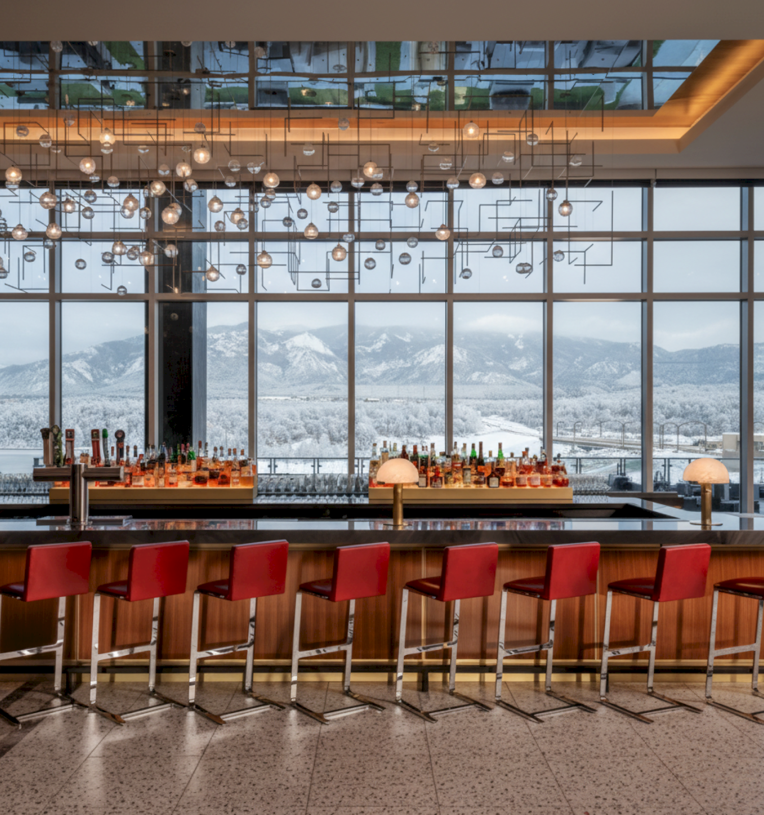 A modern bar with red stools under stylish lighting, set against large windows showing a snowy mountain landscape in the background.