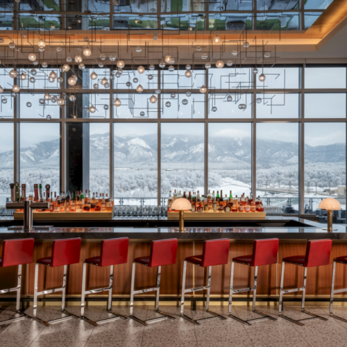 A stylish bar with red stools lining a long counter, hanging glass lights, and a snowy mountain view through large windows.