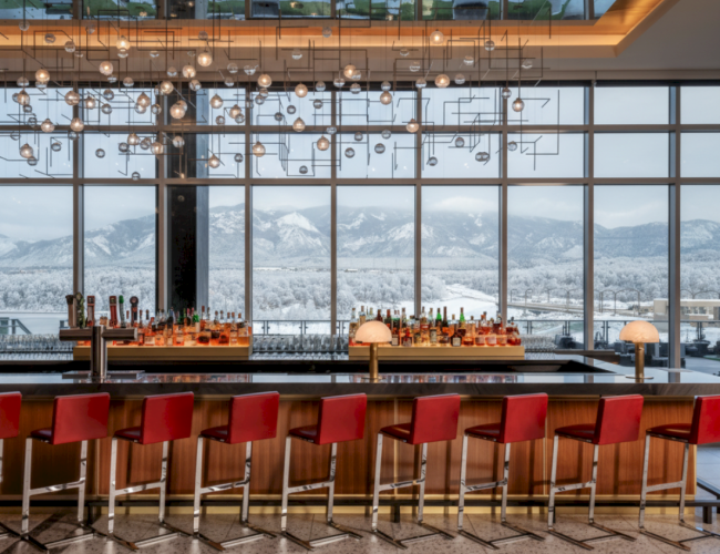A stylish bar with red stools lining a long counter, hanging glass lights, and a snowy mountain view through large windows.