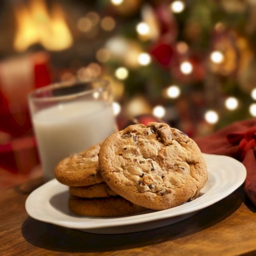 A plate of cookies with a glass of milk sits on a table in front of a festive, holiday-lit background.
