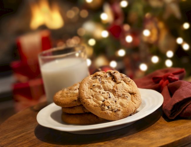 A plate of cookies with a glass of milk sits on a table in front of a festive, holiday-lit background.