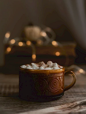 A decorative brown mug filled with marshmallows on a wooden surface, with blurred warm lights in the background creating a cozy ambiance.